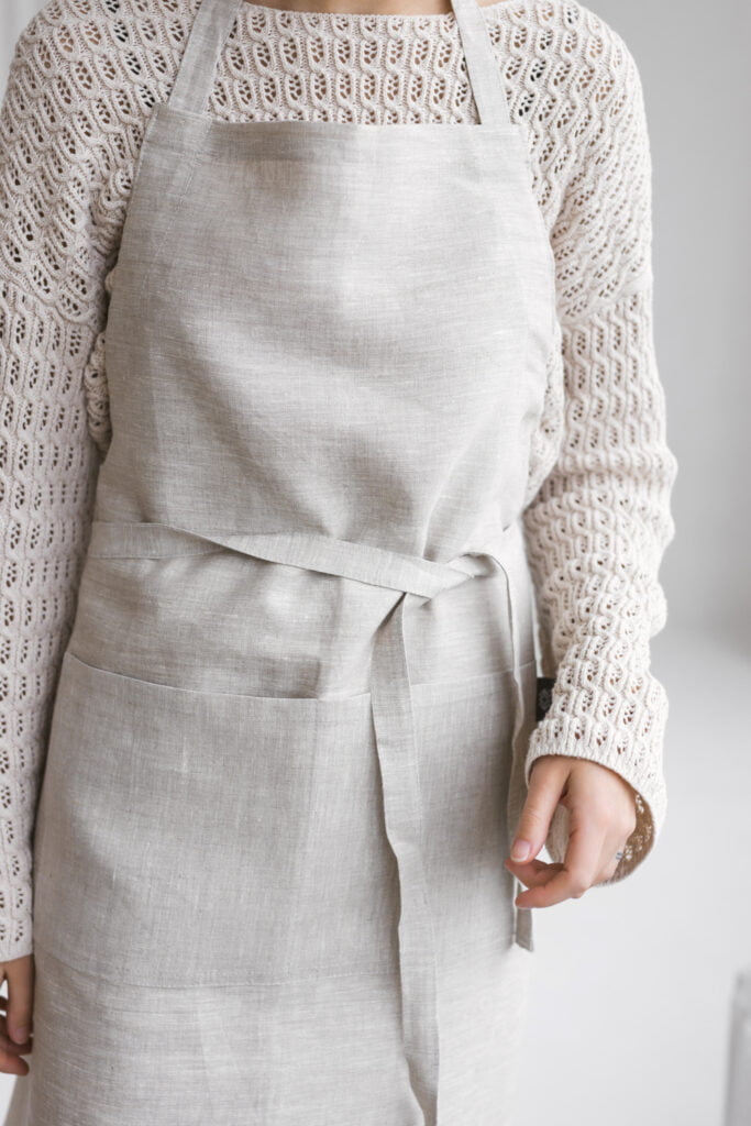 Close-up of a light gray linen bib apron showing the fabric texture, large front pocket, and knotted waist tie